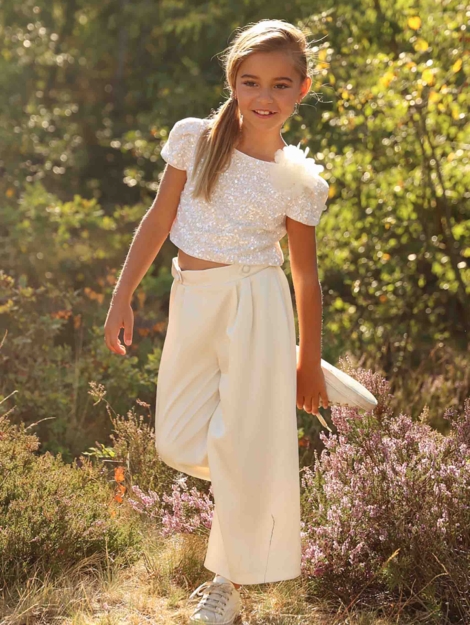 Meisje dat in de natuur staat op een been in een champagnekleurige communie outfit van Linea Raffaelli Kids - Pailletten crop top in een mooie champagne kleur met een bloem langs de halslijn. Gecombineerd met een satijnen palazzo broek met plooien en knoopjes op de tailleband. Girl standing in nature on one leg in a champagne-coloured communion outfit from Linea Raffaelli Kids - Sequin crop top in a beautiful champagne colour with a flower along the neckline. Combined with satin palazzo trousers with pleats and buttons on the waistband.