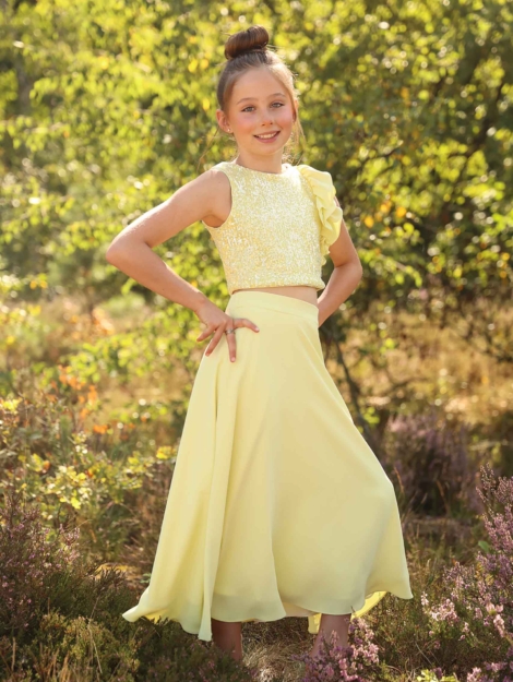 Meisje dat in de lens kijkt, in de hei gekleed in een zacht gele communie outfit van Linea Raffaelli Kids - zacht gele pailetten top met een ruche detail aan de mouw, gecombineerd met een lange, gele, soepel vallende crêpe rok. Girl that looks in to the camera dressed in a soft yellow communion outfit from Linea Raffaelli Kids - Soft yellow sequin top with a ruffled armdetail matched with a long, yellow, flowing crêpe skirt